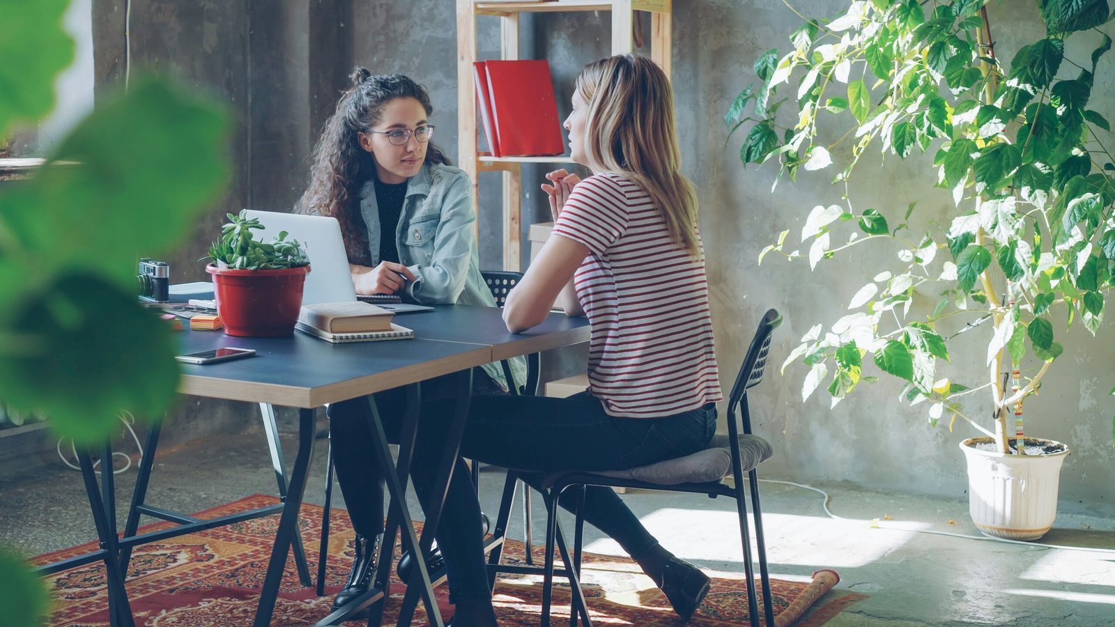 Two women are sitting at a desk, conversing.